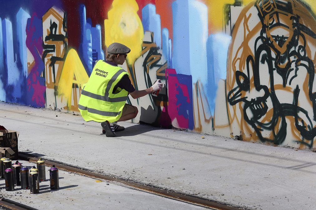 Nuori lippispäinen katutaiteilija maalaa graffitia betoniseinään.
