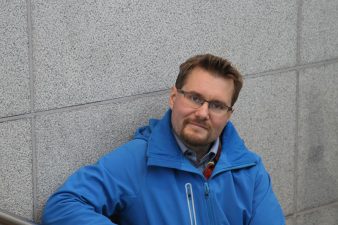 Mies seinän vieressä katsoo kameraan.