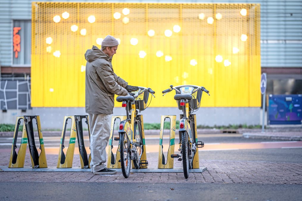 Mies seisoo pyörän vieressä kaupunkipyörätelineellä. taustalla keltainen seinä.