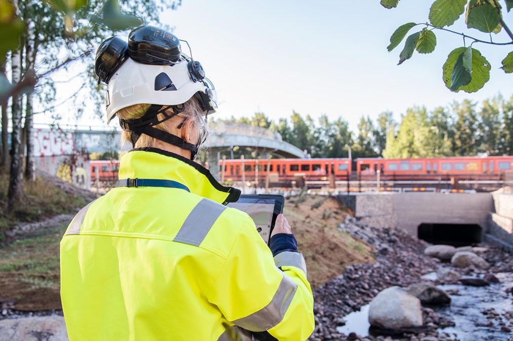 Nuori nainen huomioliivissä ja valkoisessa kypärässä ottaa kuvaa kaukana liikkuvasta metrojunasta.