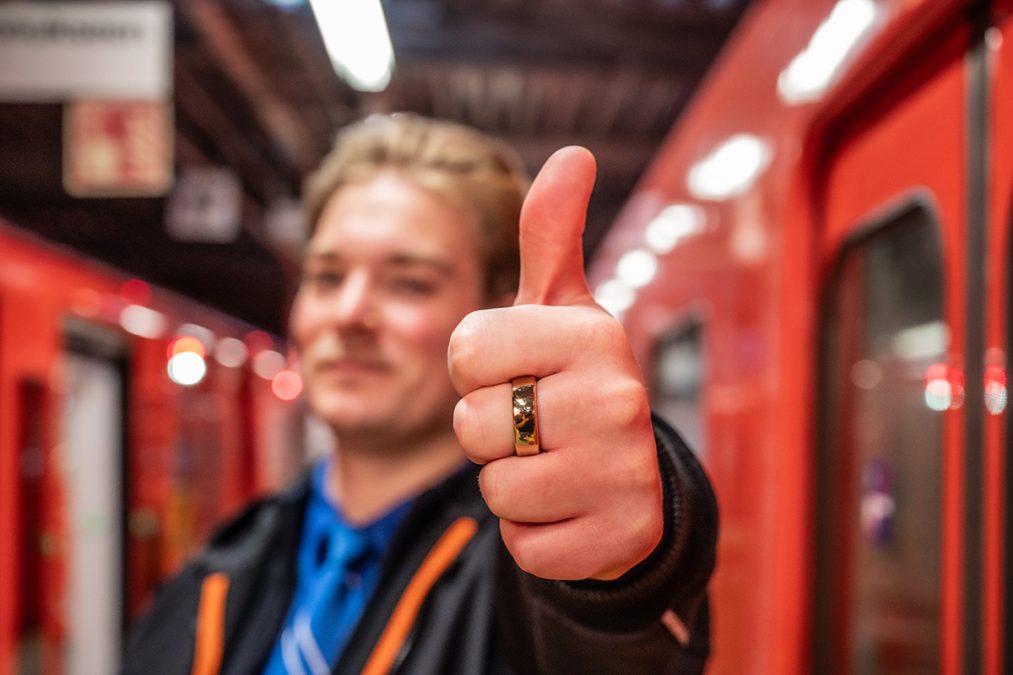 Henkilö pitää peukaloa ylöspäin, taustalla on metrojuna.