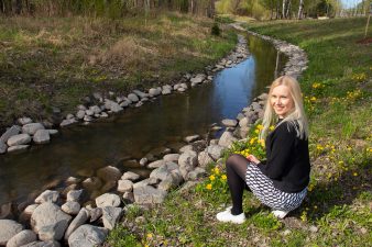 Vaalea nainen istuu niityllä veden ääressä.