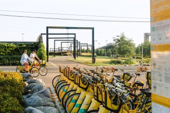 Kaksi henkilöä pyöräilee keltaisilla kaupunkipyörillä. Edessä kaupunkipyöräteline.