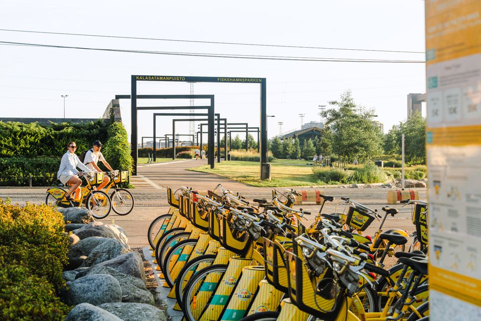 Kaksi henkilöä pyöräilee keltaisilla kaupunkipyörillä. Edessä kaupunkipyöräteline.