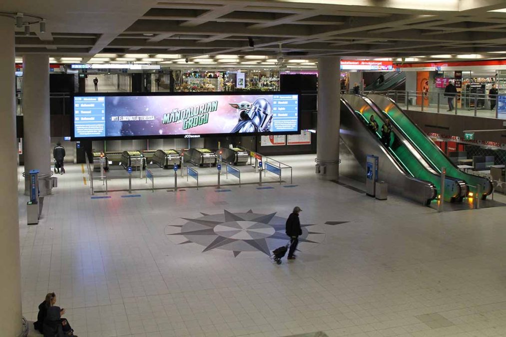 Rautatientorin metroaseman kompassitasolla ihmisiä, taustalla liukuportaat ja mainosnäyttö.
